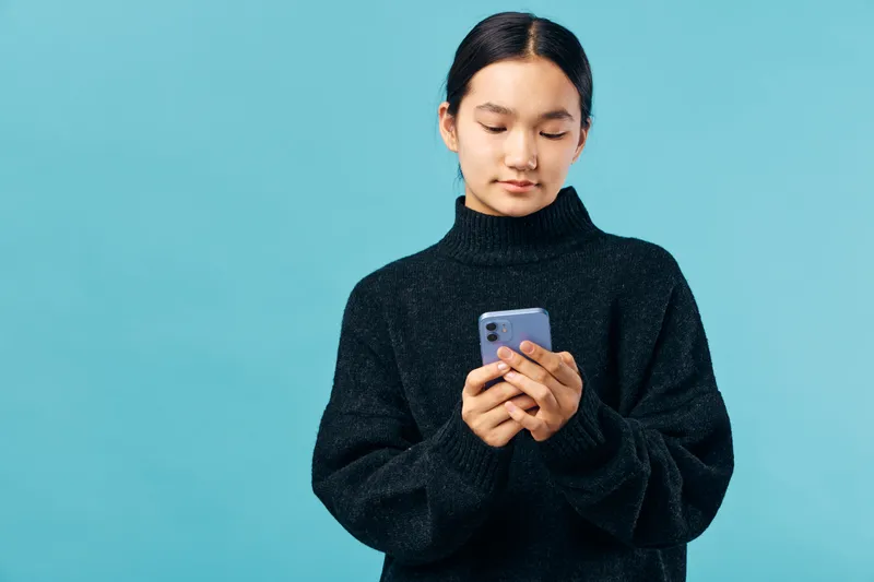 Young woman in black sweater looking at smartphone on blue background. Casual person texting or browsing on mobile phone indoors with focus on device and hands in portrait.
