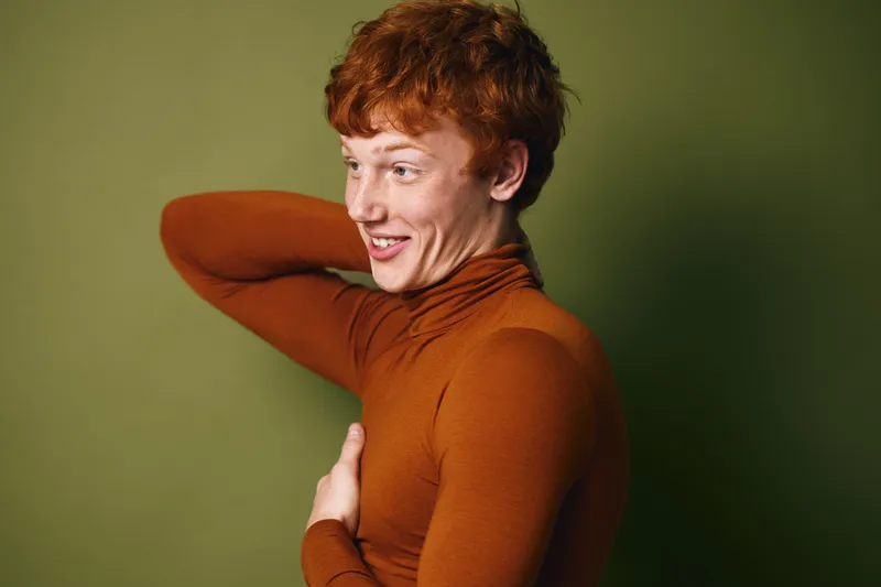 Young man with red hair and freckles wearing a brown turtleneck sweater posing with hand behind neck and smiling on green background in studio portrait.