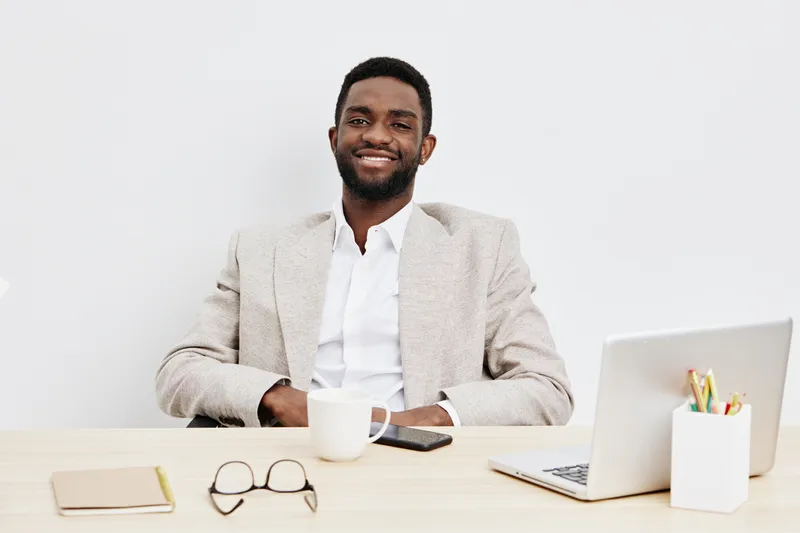 Smiling man businessman in beige blazer and white shirt sitting at desk with laptop, coffee mug, glasses, sketchpad, and pen on plain white background. Business lifestyle concept.