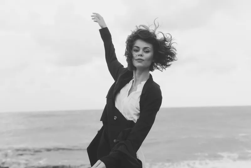Woman in a blazer and white shirt stands by the sea, wind tousling hair, confident pose, outdoor fashion, dynamic movement and moody shoreline atmosphere