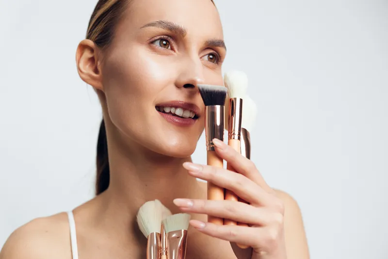 Woman applying foundation with brush near her cheek, closeup portrait indoors, beauty routine and skincare, holding makeup brushes, studio soft lighting, warm smile, selfcare concept