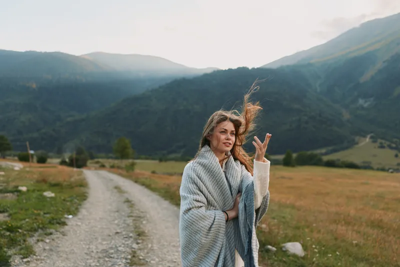 A woman wrapped in a cozy blanket stands on a rural dirt road surrounded by mountains and fields, enjoying a peaceful moment in nature during a cool day.