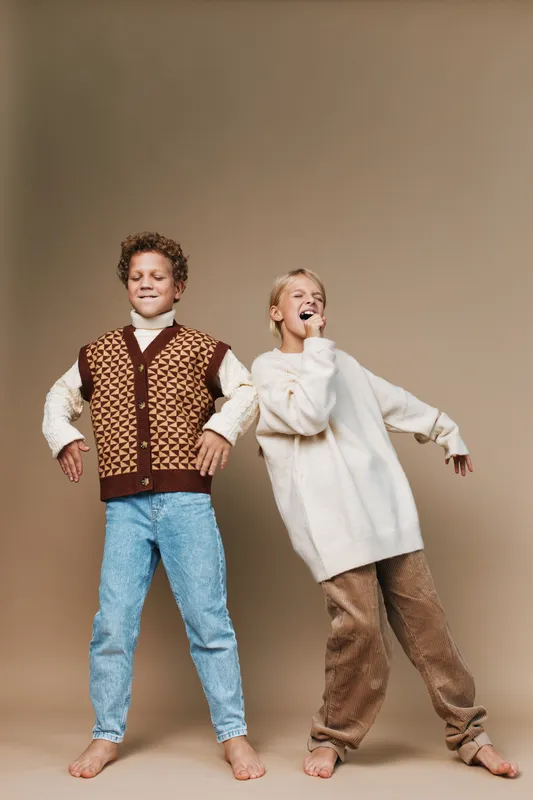 Two cheerful children pose playfully in cozy outfits, one wearing a patterned knit vest with jeans and the other in an oversized sweater with loose pants, smiling confidently.