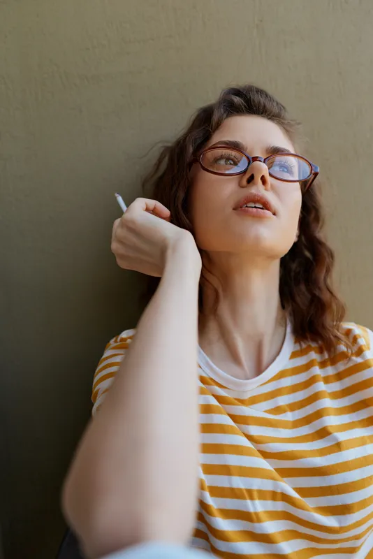 Young woman smokes a cigarette while leaning against a plain wall in a casual indoor setting, conveying a thoughtful mood with a relaxed posture.
