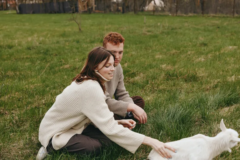 A couple sits in a sunlit field, smiling as a goat kid approaches, sharing a moment of lifestyle and companionship with nature and pasture scenery.
