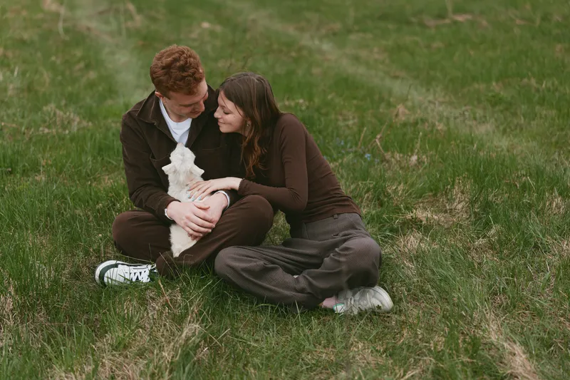 Happy couple in casual clothes cuddle a goat kid on a green hillside, sharing a tender outdoor moment in nature with calm grass, gentle sunlight, and a warm lifestyle vibe.