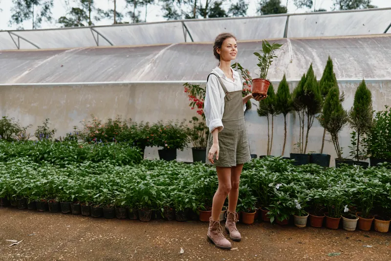 Young woman in a gardening outfit holding a potted plant, surrounded by greenery in a greenhouse, exuding joy and passion for gardening