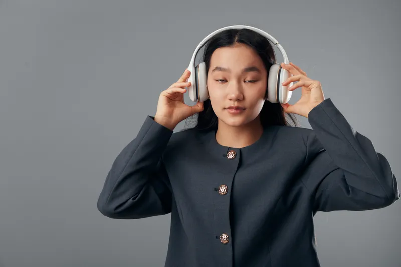 Woman with headphones on head standing against grey background adjusting audio device with closed eyes in casual stylish jacket for listening relaxation and music enjoyment.