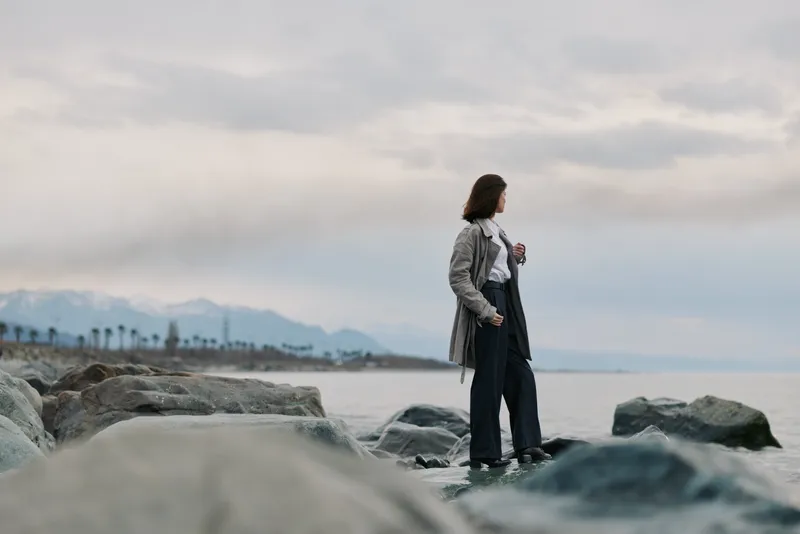 Woman in casual clothes stands on rocky shore looking at cloudy sky over calm sea. Concept of solitude, contemplation, and nature connection during overcast weather outdoors.