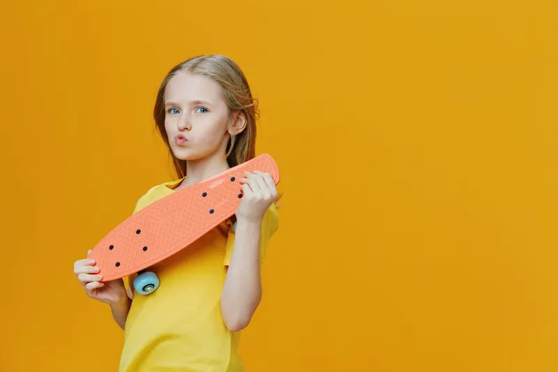 Young girl with blue eyes holding an orange skateboard, wearing a yellow shirt, against a bright yellow background, conveying a playful and vibrant mood