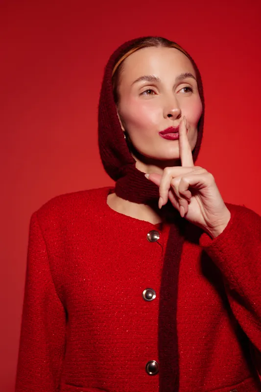 Woman in red coat and headscarf makes silence gesture with finger on lips, looking aside on red background. Concept of secrecy, quiet, and calm expression in fashion portrait.