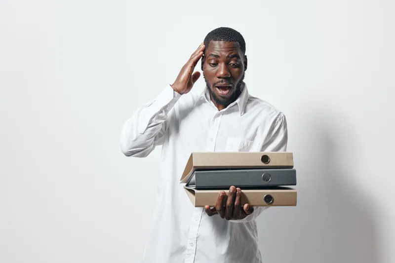 African American man in white shirt holding folders with surprised expression, standing against white background. Office worker with dark skin managing documents and files.