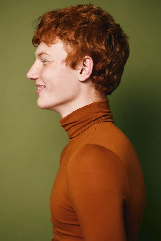 Young man with red hair and fair skin in orange turtleneck smiling in profile against green background, casual fashion portrait, studio lighting and confident expression.