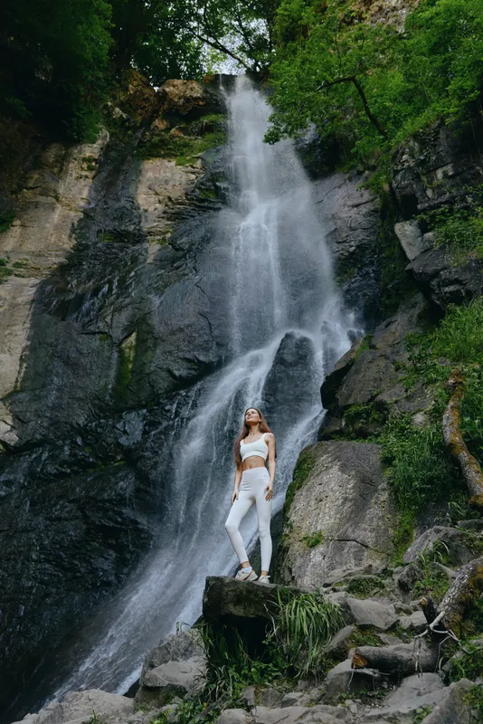young woman standing at the base of a tall waterfall surrounded by green foliage and rocky cliffs, wearing white clothes and looking upward in nature