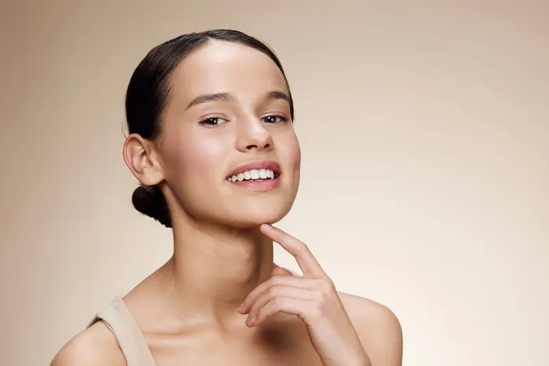 Young woman smiling softly with a natural look, showcasing healthy skin and positivity against a soft beige background, ideal for wellness and beauty concepts
