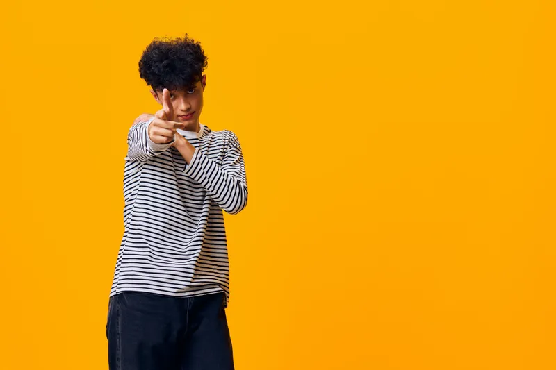 teenage man pointing finger playfully on vibrant orange background wearing striped shirt and dark pants with casual confident pose and curly hair