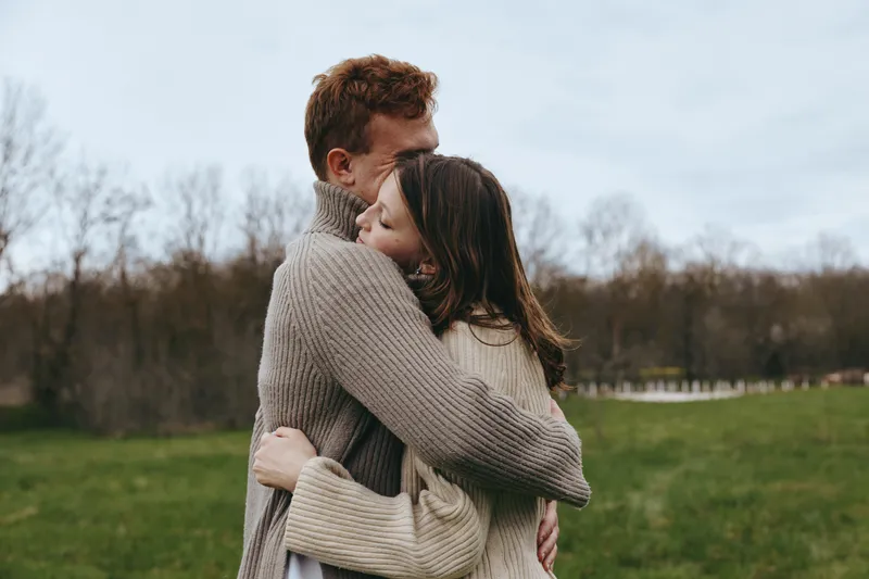 Couple embrace outdoors in cozy sweaters, showing tenderness, love, and connection in a calm countryside setting, lifestyle moment with soft light and natural colors, intimate mood and warm