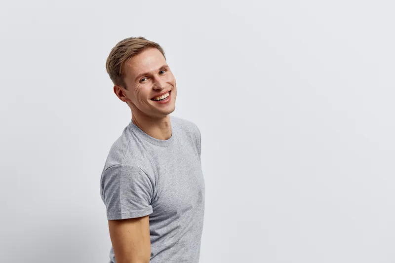 Happy smiling young Caucasian man with light brown hair wearing a casual gray t-shirt standing isolated on white background studio portrait. People lifestyle concept, positive emotion, relaxed mood