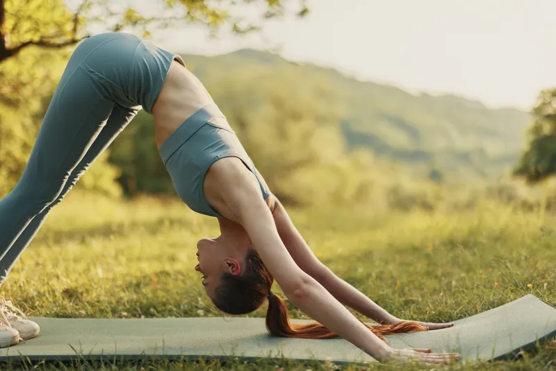 Young woman practicing yoga in downward dog pose outdoors, surrounded by nature, conveying serenity and health in a sunny setting