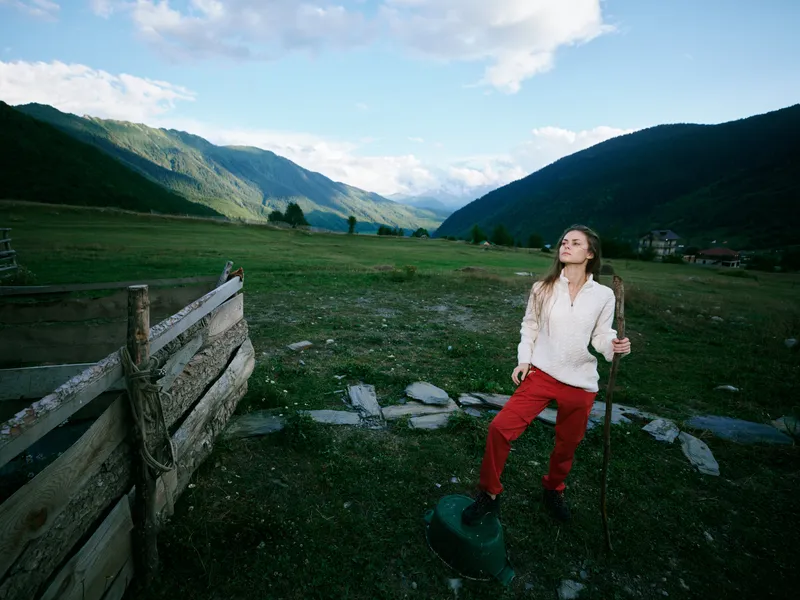 Hiking, mountains, landscape, outdoors, trekking, valley, scenery, adventure, a woman leans on a trekking pole beside a rustic fence in a wide alpine meadow under blue skies