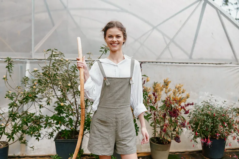 Young woman in casual gardening attire smiling in a greenhouse setting, surrounded by lush plants and greenery, ideal for themes of gardening and healthy living