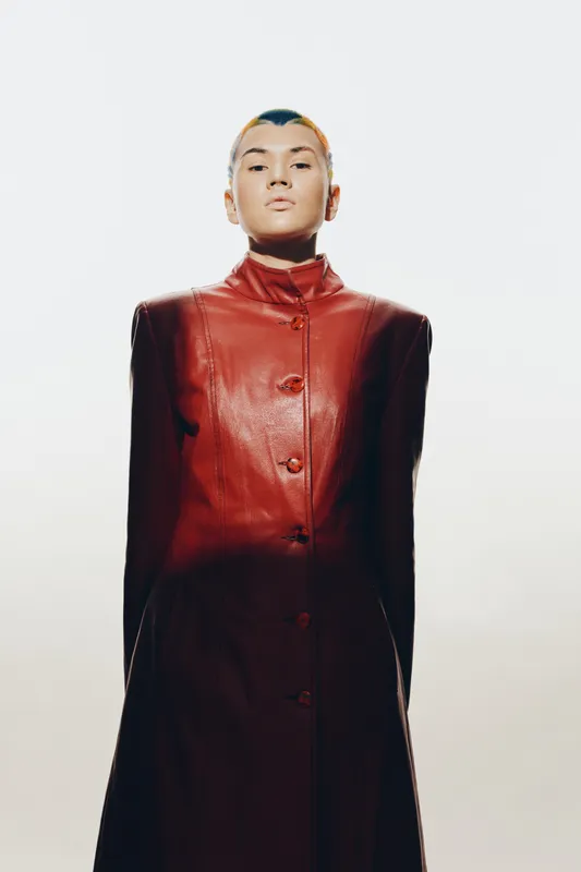 young woman in a red leather coat standing confidently against a light background, modern fashion style, minimalistic portrait with strong posture