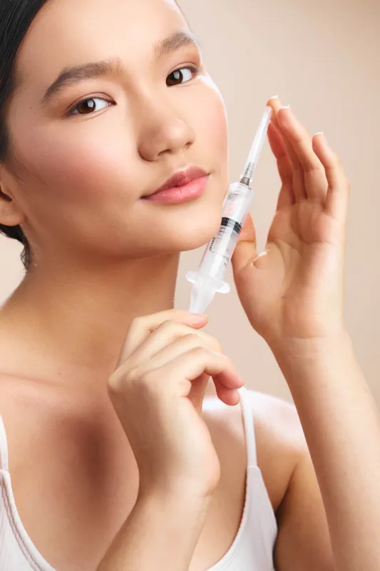 Asian woman prepares a syringe in a clinical setting, with a calm expression and steady hands, conveying care, focus, and precision in a medical environment.