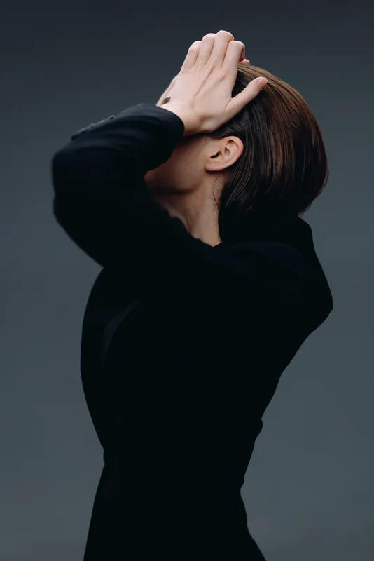 Side view of a woman in black clothing covering her face with hands, moody atmosphere, studio background, artistic expression