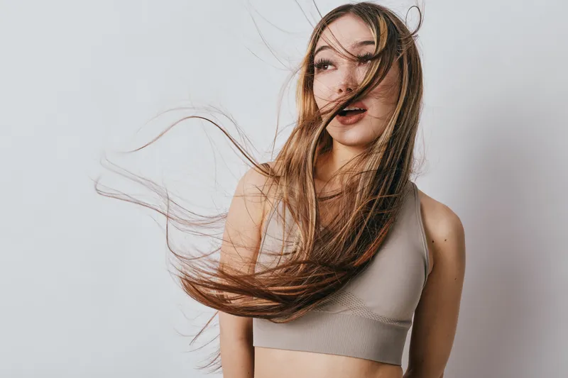 Woman with long hair blowing in the wind wearing sleeveless top standing against light background. Beauty, motion, hair movement and casual style portrait of young female.