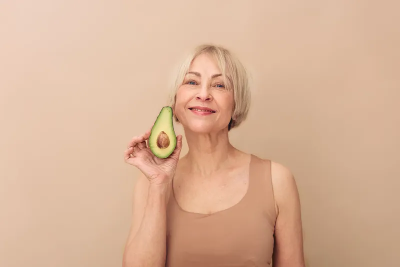 Smiling mature woman holds fresh avocado near face on beige background. Healthy lifestyle concept with natural food, nutrition, and positive wellbeing for older adults and beauty care.
