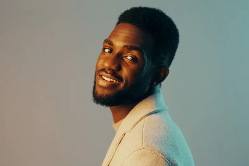 A cheerful Black man in a stylish light sweater smiles confidently against a soft gradient background, embodying warmth and creativity in a modern portrait