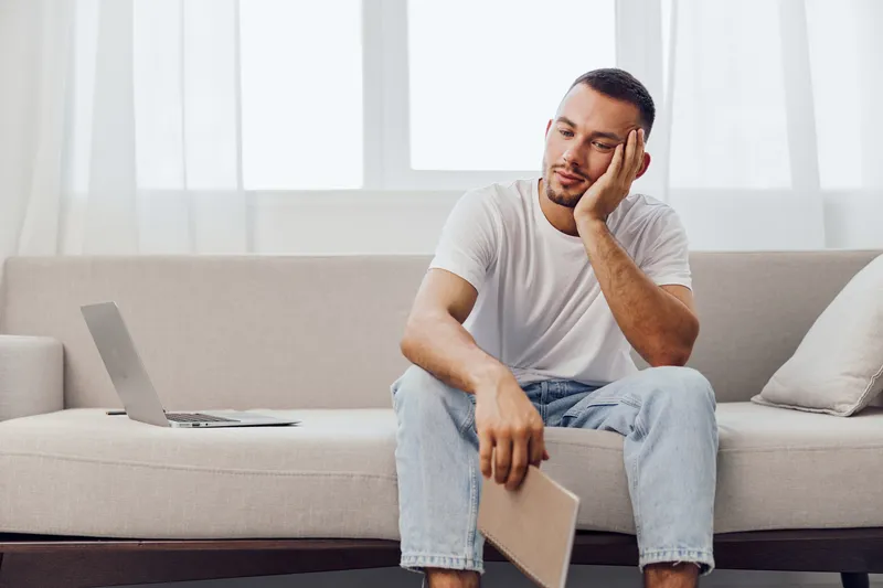 Pensive man sitting on a couch, reflecting on work-life balance and digital detox, portraying anxiety and emotional minimalism in a serene home environment.
