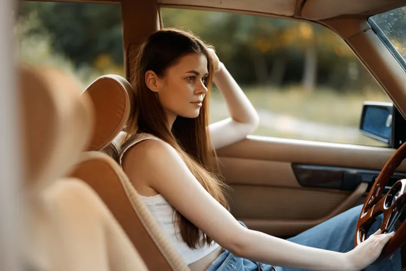 Young woman with long hair sitting in a vintage car, wearing casual clothes, relaxed expression, and enjoying the outdoor scenery during a sunny day.