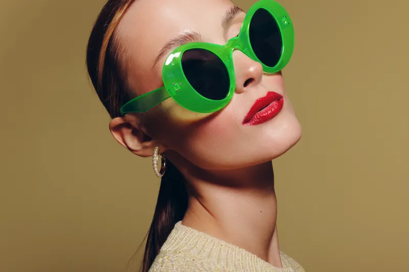 Stylish woman with green sunglasses and red lipstick poses against beige background. Fashion portrait with trendy accessories and confident expression in studio lighting.