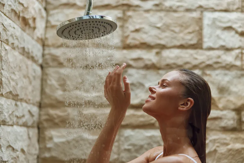 woman enjoying outdoor shower with falling water against natural stone wall background, relaxed expression, wellness and self-care moment