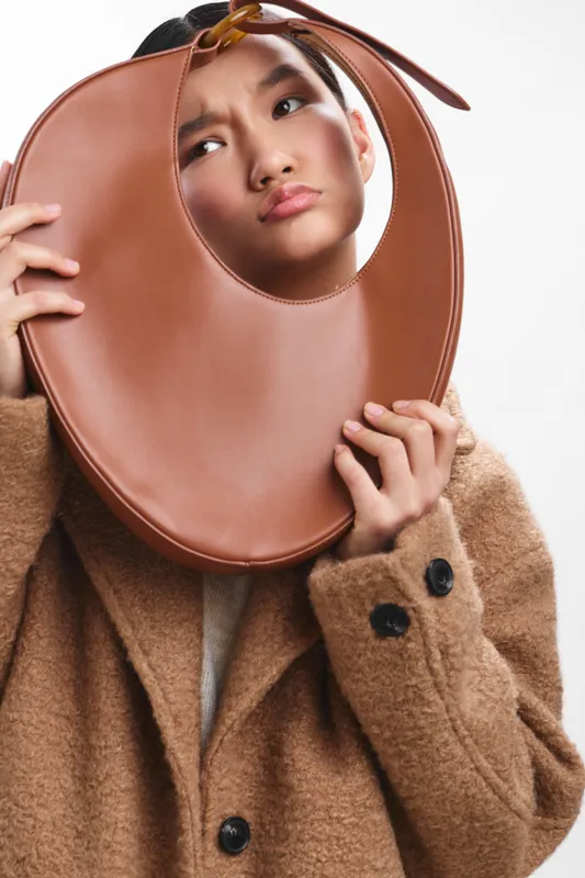 Asian woman holds a large circular brown accessory near her face, posing in a studio with soft lighting and minimalist backdrop, creating a stylish, thoughtful, and contemporary mood.