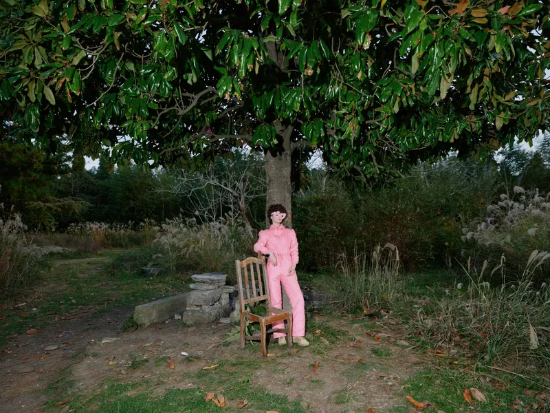 Outdoor garden scene with a woman in a pink suit standing beside a wooden chair under a large tree, capturing a calm portrait amidst nature and rustic surroundings in soft daylight.