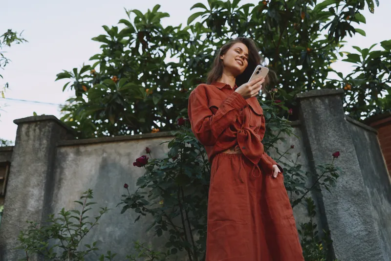 A smiling woman in a red dress uses her phone outdoors in a garden, showcasing a casual lifestyle moment and relaxed confidence, natural light, candid mood, everyday fashion