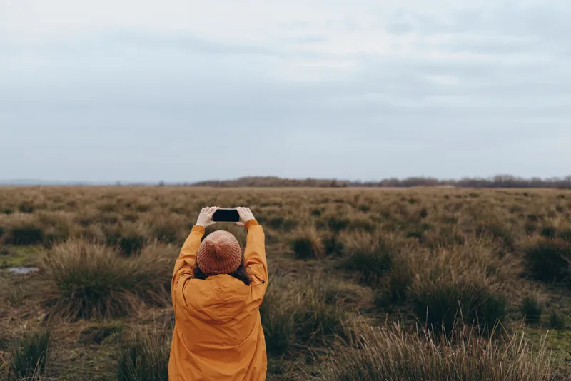 Woman in yellow hooded jacket stands in a grassy field from behind, hands on head. Outdoors nature lifestyle with calm autumn landscape, soft cloudy sky. Cold wind brings misty calm.