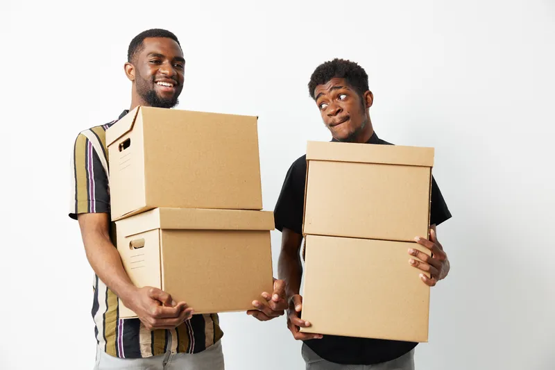 Two smiling African American men are holding cardboard boxes, showcasing teamwork and positivity in a bright, minimalistic environment. Their cheerful expressions enhance the scene.