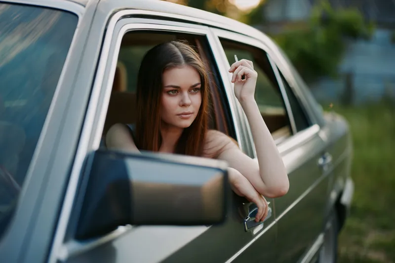 Young woman with long hair leaning out of a vintage car window, exuding a contemplative mood while holding a cigarette, surrounded by nature during golden hour.