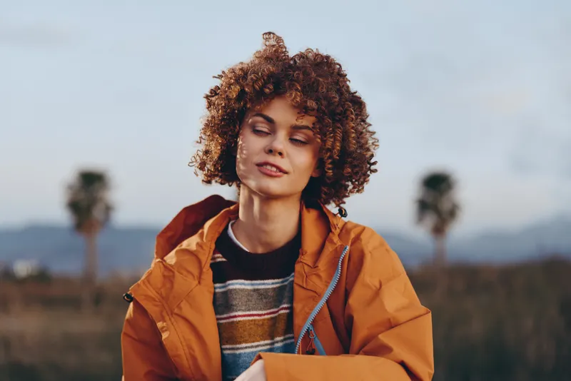 Woman lifestyle outdoors, curly hair and orange jacket, standing on a rural field at dusk, soft sunlight, relaxed gaze toward camera, calm mood, distant mountains blur in background