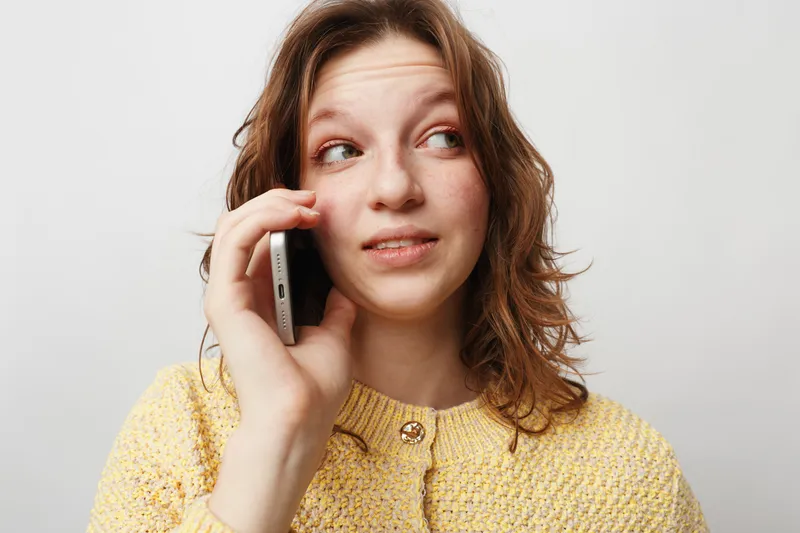Young woman with curly hair wearing yellow sweater talks on smartphone, looking sideways with thoughtful expression. Isolated on white background, concept of communication and casual lifestyle.