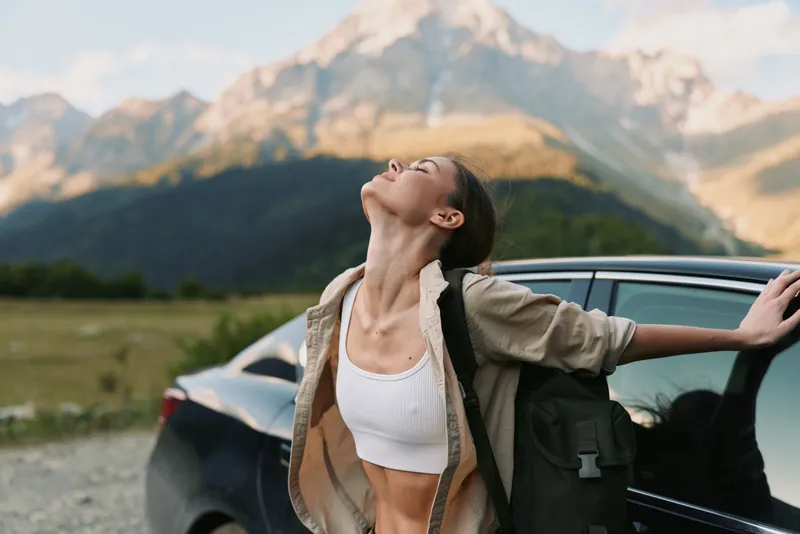 Young woman enjoys fresh air after a hike, leaning against a dark car with distant rugged mountains, blue sky and open countryside behind, feeling liberated and alive