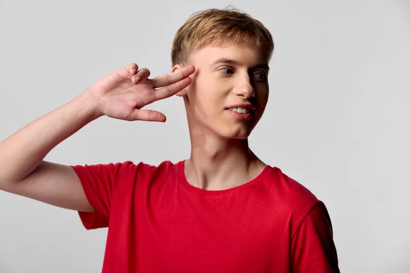 Listening gesture, ear close up, young man in a casual red shirt signals attentive communication, playful pose highlighting focus, awareness and listening skills for education or stock imagery about