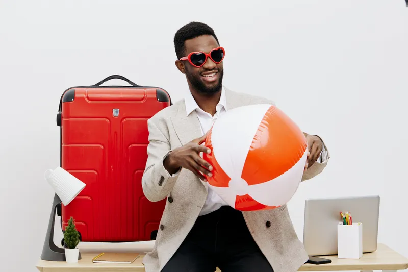 Happy joyful man with dark skin wearing sunglasses holding beach ball, standing in modern office with travel accessories like suitcase and laptop, preparing for vacation indoor. People lifestyle and t