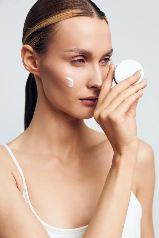 Young woman applying cream on face, skincare routine, natural beauty, clean skin, minimal makeup, white background, focused expression, healthy lifestyle, moisturizing product