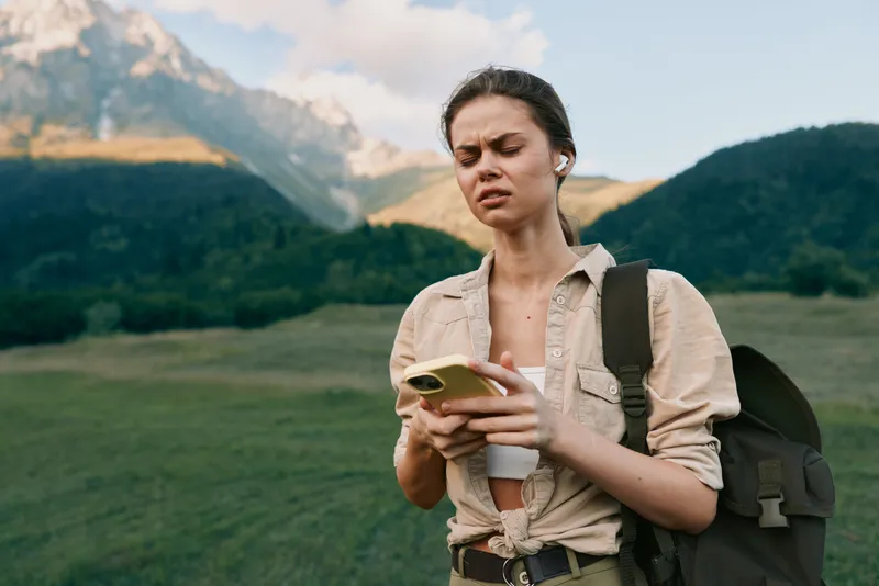 A mountain landscape scene shows a young woman traveler reading on a smartphone outdoors, wearing a beige shirt and backpack, deep in concentration, exploring with curiosity.