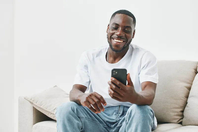Happy African American man smiling while using smartphone at home, wearing casual white t shirt and jeans, conveying joy and comfort in a bright setting