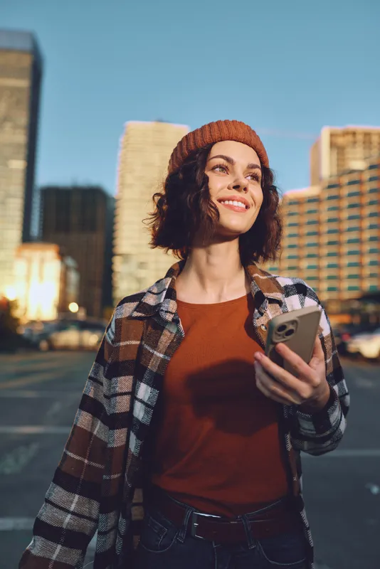 woman urban smartphone smile candid lifestyle authenticity golden glow mindful living emotional storytelling. Young woman in plaid and beanie walks city street at golden hour holding phone, radiant
